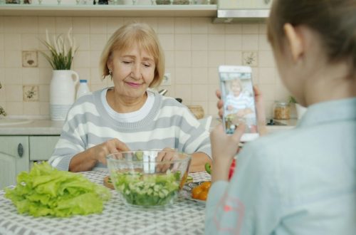 Little girl is taking pictures of granny who is cooking salad then posing for camera with cucumber slices having fun, child is using modern smartphone camera.