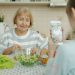 Little girl is taking pictures of granny who is cooking salad then posing for camera with cucumber slices having fun, child is using modern smartphone camera.